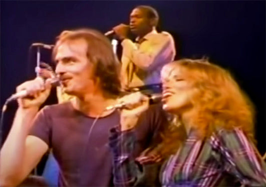 Live performance by two musicians singing on stage during a concert. One man with long hair is holding a microphone, while a woman with curly hair and a striped shirt sings alongside him. Another performer can be seen in the background.