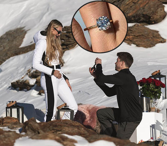 Man proposing to his girlfriend on a snowy mountain, kneeling while offering an engagement ring. She stands smiling, wearing a white ski outfit. An inset shows a close-up of the stunning blue gemstone ring. The scene is decorated with roses and lanterns.
