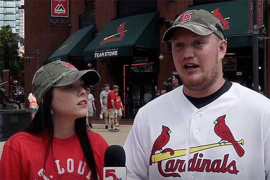 Two St. Louis Cardinals fans, a man in a jersey and a woman in a red shirt, standing outside the Cardinals team store, speaking into a microphone while wearing matching green baseball caps.