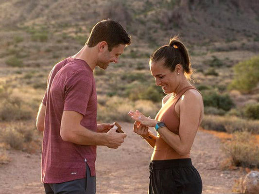 A couple enjoying an outdoor engagement, with the man holding a small box while the woman admires a ring on her finger, set against a beautiful desert landscape during golden hour.