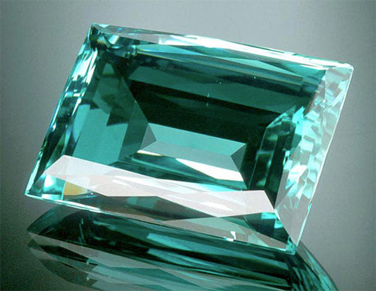 A large, emerald-cut aquamarine gemstone, showing brilliant clarity and vivid blue-green color, displayed on a reflective surface.