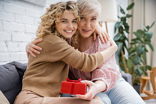 Mother and daughter hugging each other while holding a red gift box, smiling and enjoying a joyful moment together in a cozy living room.