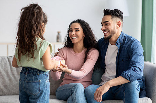 Happy family of three with a young girl giving a gift box to her smiling parents. The father and mother are seated on a sofa, enjoying the moment in a cozy living room.