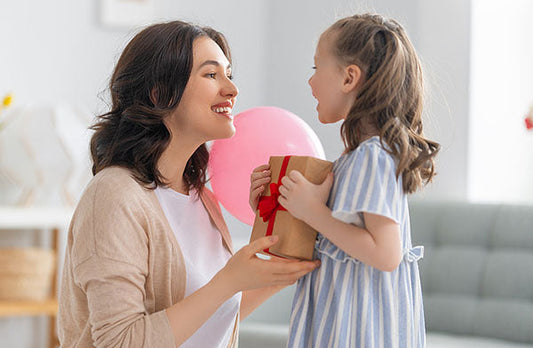 Mother and daughter smiling at each other, with the daughter holding a gift wrapped with a red ribbon, and a pink balloon in the background.