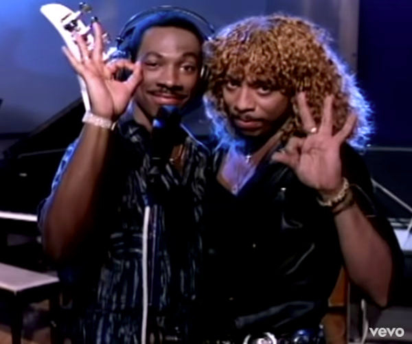 Two men posing together in a recording studio, both flashing a hand gesture, with one wearing a curly wig and the other holding a saxophone, reflecting an iconic 1980s music vibe.