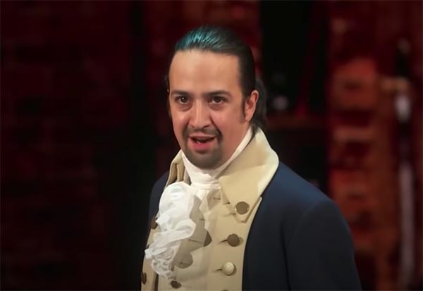 Lin-Manuel Miranda performing as Alexander Hamilton in the Broadway musical 'Hamilton', dressed in a historical costume with a white cravat and jacket, delivering an intense expression.