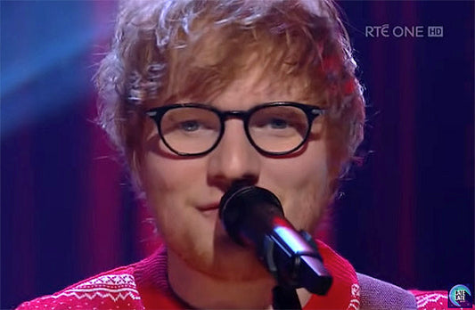 Ed Sheeran performing on stage with a guitar, wearing glasses and a red festive sweater, smiling while singing into a microphone.