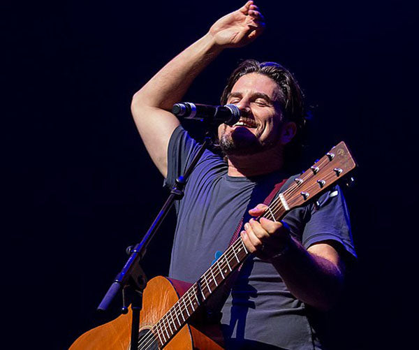 Smiling musician performing on stage with a guitar, holding a microphone and raising one arm in excitement, with a dark background and stage lights.