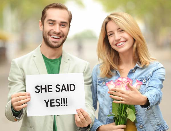 Happy engaged couple celebrating, man holding a sign that reads 'SHE SAID YES!!!' while the woman holds a bouquet of pink roses, both smiling outdoors