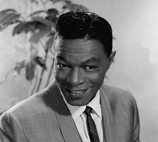Black and white portrait of Nat King Cole, a famous jazz musician, smiling while wearing a suit and tie, with a plant in the background.