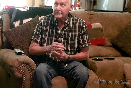 Older man sitting on a couch with a plaid shirt, holding a small object, and a remote control on the side table.