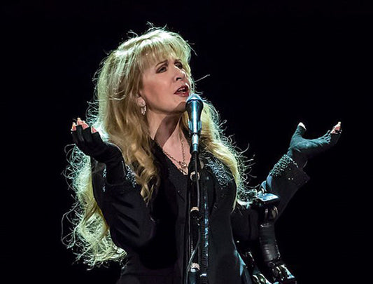 Stevie Nicks performing on stage with dramatic lighting, wearing a black outfit and gloves, showcasing her signature style and stage presence.