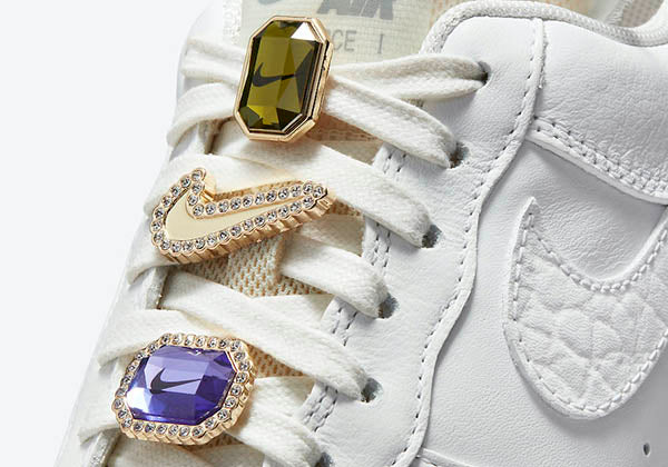 Close-up of white Nike Air Force 1 sneakers with jeweled lace charms featuring purple, green, and clear stones, highlighting the iconic Nike logo.