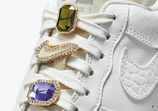 Close-up of white Nike Air Force 1 sneakers with jeweled lace charms featuring purple, green, and clear stones, highlighting the iconic Nike logo.