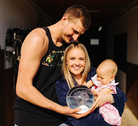 Nikola Jokić, the NBA player, poses with his wife and baby, holding an award. The family smiles together, with Jokić dressed casually in a tank top and his wife and child dressed in casual attire.