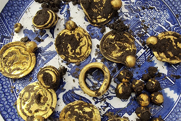 Ancient gold coins and a ring partially covered in dirt, displayed on a blue patterned plate, representing a treasure discovery.