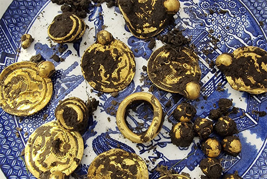 Ancient gold coins and a ring partially covered in dirt, displayed on a blue patterned plate, representing a treasure discovery.
