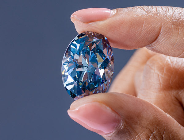 Close-up of a hand holding a large, stunning blue diamond with intricate facets, highlighting the diamond's clarity and brilliance.