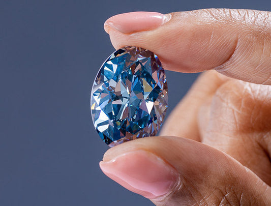 Close-up of a hand holding a large, stunning blue diamond with intricate facets, highlighting the diamond's clarity and brilliance.