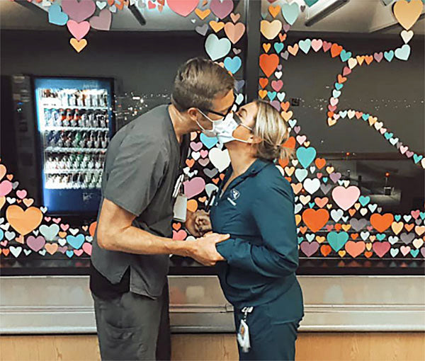 Two healthcare workers in masks sharing a kiss in front of a window decorated with colorful heart-shaped paper cutouts.