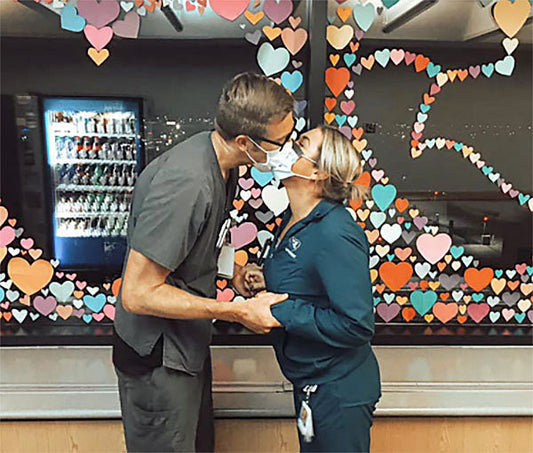 Two healthcare workers in masks sharing a kiss in front of a window decorated with colorful heart-shaped paper cutouts.