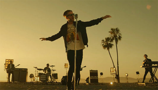 Musician performing on a beach at sunset with a microphone stand, while the band plays in the background, illuminated by stage lights.