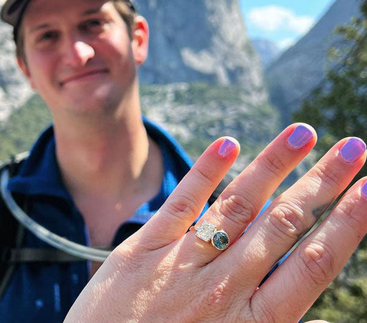 Engaged couple outdoors with the woman showing off her engagement ring featuring a large diamond and a green gemstone, while the man smiles in the background, surrounded by a scenic mountain view.