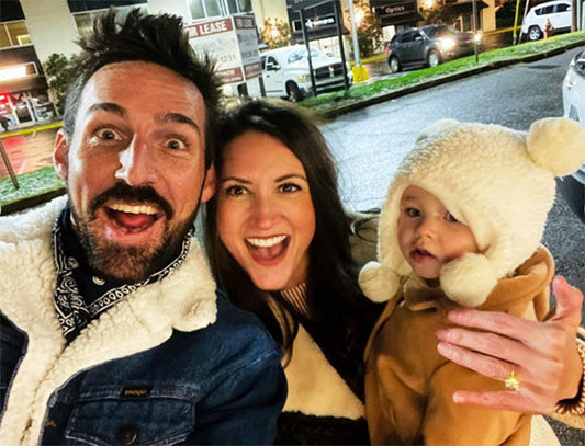 A happy family of three taking a selfie outdoors, with the mother, father, and baby wearing cozy winter attire, smiling at the camera with a festive background.
