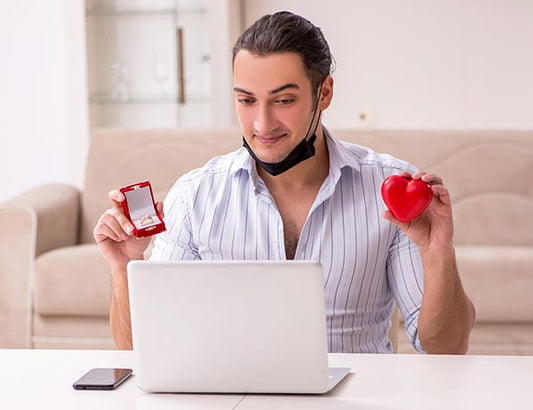 A man sitting at a laptop, holding a red heart-shaped box in one hand and a red jewelry box containing an engagement ring in the other, smiling as he prepares to propose.