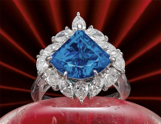 A luxurious blue sapphire ring with a diamond halo, featuring a large triangular-shaped sapphire surrounded by marquise-cut diamonds, set against a red radial background.
