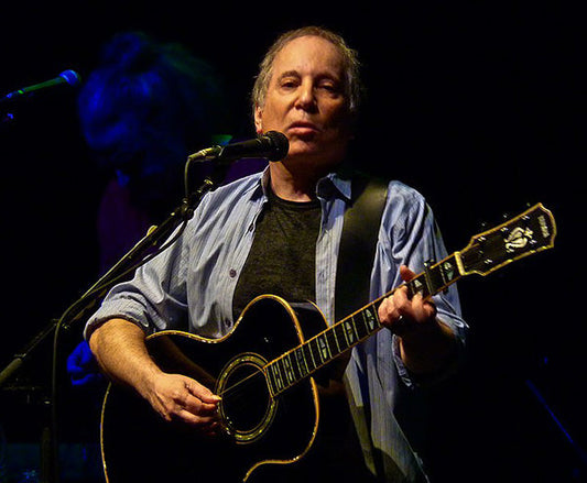 Paul Simon performing on stage with an acoustic guitar during a concert, wearing a blue shirt and singing into a microphone, illuminated by stage lights.