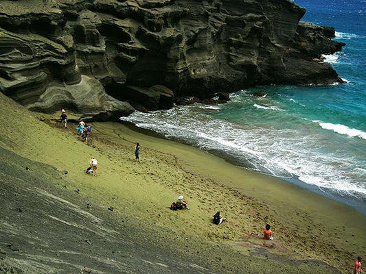 People walking and relaxing on a green sand beach near rocky cliffs with blue ocean waves.