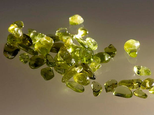 Pile of peridot gemstones, small and translucent, scattered on a reflective surface with a green hue, glistening in the light.