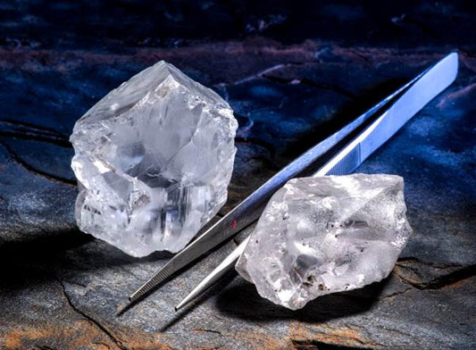 Close-up of rough clear quartz crystals being held by a pair of tweezers, set against a dark stone surface. The image highlights the transparency and natural facets of the crystals.