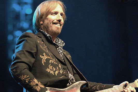 Tom Petty performing on stage with a guitar, smiling while wearing a black embroidered jacket, captured during a live concert.