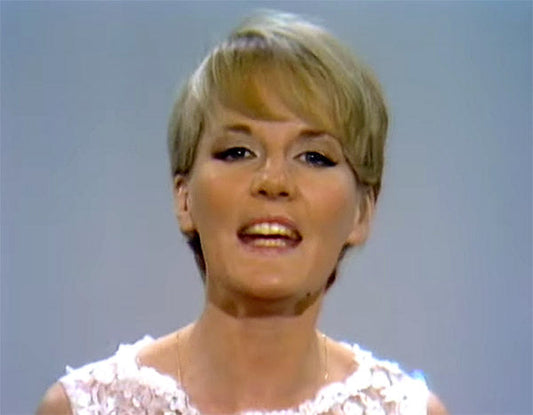 Black and white image of a woman with short blonde hair, wearing a white lace top, singing and speaking on stage.