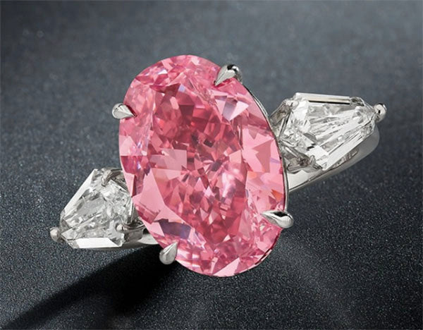 A close-up of an elegant pink oval-shaped gemstone ring with two white diamond side stones, set in a silver band, showcasing a luxurious and vibrant design.