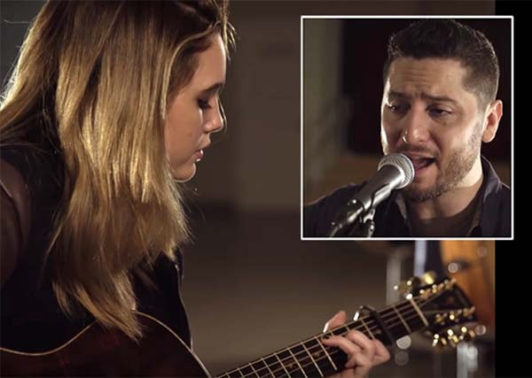Female musician playing acoustic guitar in a recording studio while a male singer performs into a microphone in the background. Both artists are engaged in a musical duet, captured in a close-up shot of their performance.