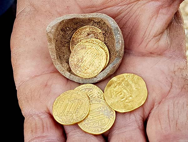 Close-up of a hand holding a stone container with several ancient gold coins resting on the palm, showcasing the shiny, detailed surface of the coins.