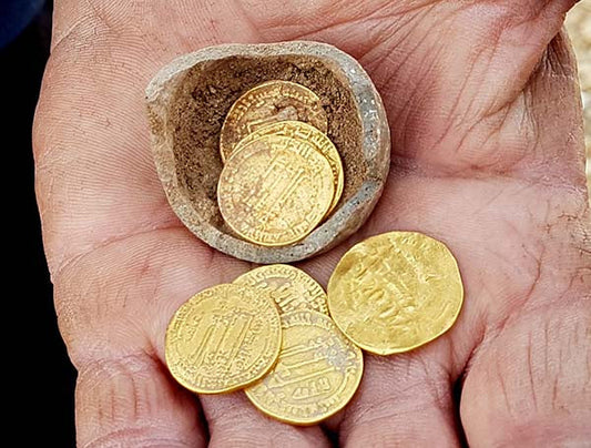 Close-up of a hand holding a stone container with several ancient gold coins resting on the palm, showcasing the shiny, detailed surface of the coins.