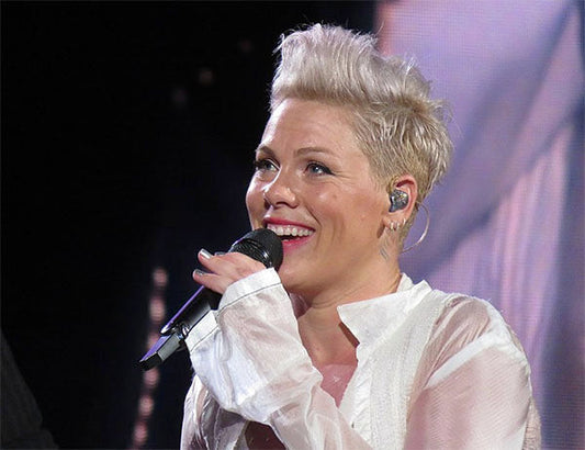 Pop singer Pink performing on stage with a microphone, smiling and wearing a white sheer blouse. Her platinum blonde hair is styled in a short, spiked cut, and she is wearing in-ear monitors during the live performance.