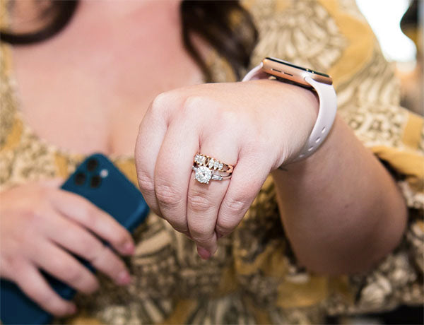 Close-up of a hand wearing a diamond engagement ring with two wedding bands, alongside a smartphone and smartwatch.