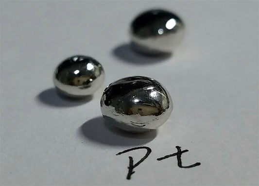 Close-up image of platinum (Pt) metal droplets on paper, with 'Pt' written next to them, showcasing the shiny metallic texture of the precious metal.