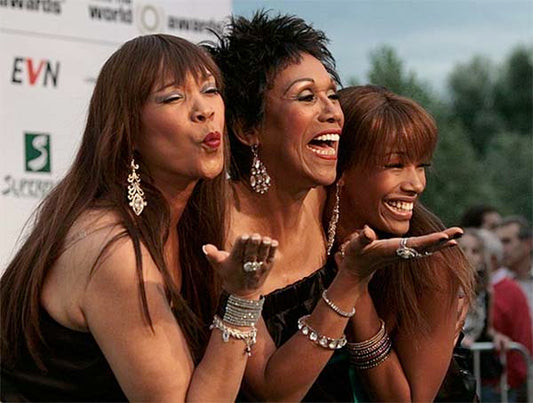Three women blowing kisses to the camera while posing together at an event, dressed in elegant black outfits with jewelry, smiling and exuding confidence.