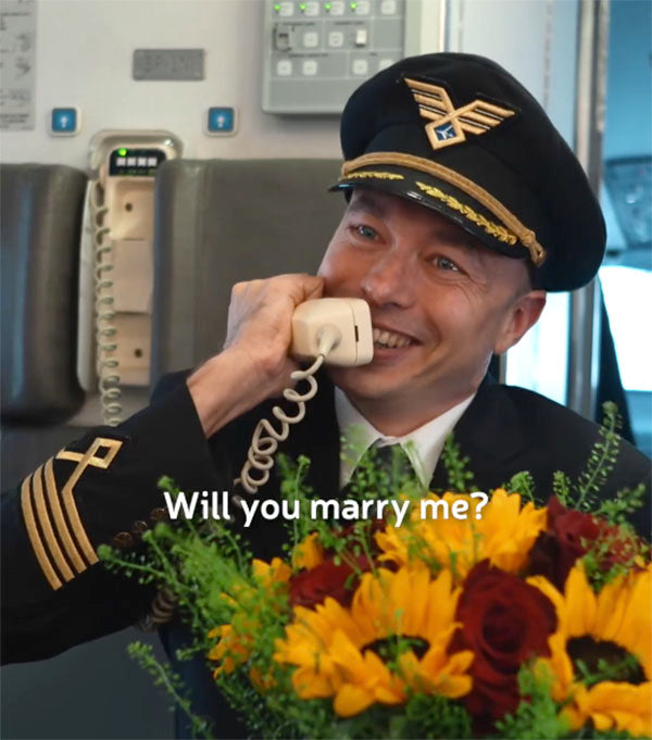 Pilot holding a bouquet of sunflowers, smiling while holding a phone, proposing marriage with the text 'Will you marry me?' displayed on the image.