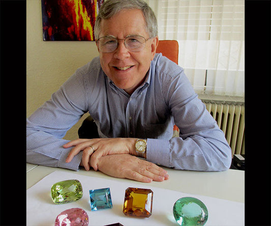 A man smiling and posing with an assortment of colorful gemstones, including green, blue, and yellow stones, laid out on a table in front of him.