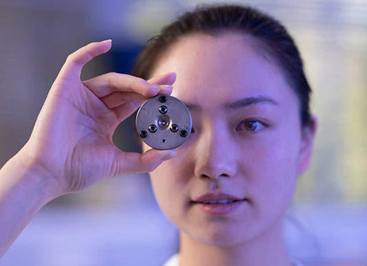 Female scientist holding a precision mechanical component with six screws, demonstrating a research tool or equipment.
