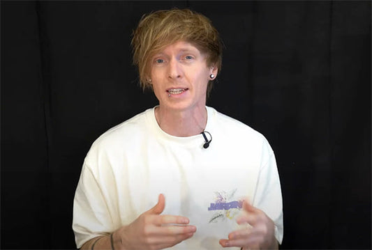 Man with light blonde hair wearing a white t-shirt, speaking and gesturing while recording a video, with a black background.