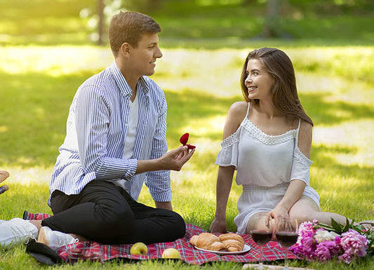 Couple enjoying a romantic picnic outdoors, with the man holding a red rose and offering it to the woman, who smiles back. They are sitting on a blanket with croissants, fruits, and flowers, surrounded by green grass in a serene park setting.