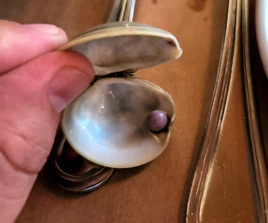 Close-up image of a hand opening an oyster shell to reveal a purple pearl inside, placed on a table with a fork and knife.
